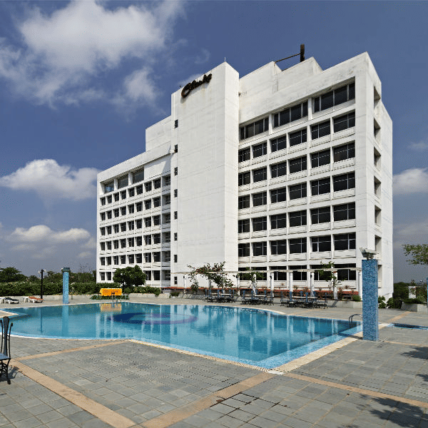 A large, modern white hotel building is shown behind a spacious blue swimming pool with lounge chairs on the patio - Clarks Avadh, Lucknow