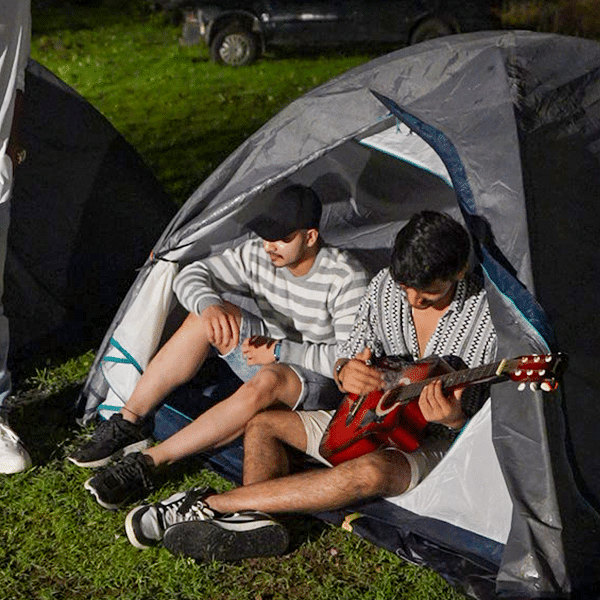 Two men sitting in a tent playing guitar at night during a camping trip | Diamond Parks, Pune.