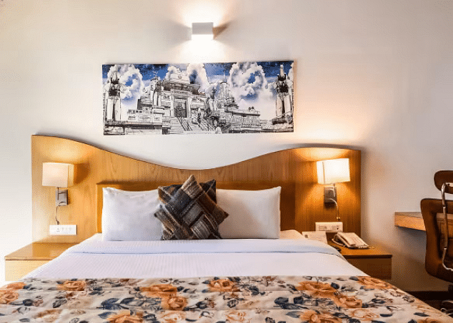 A bright room with a bed featuring a floral patterned runner, a desk, and artwork on the wall. - Bamboo Saa Resort & Spa, Udaipur.