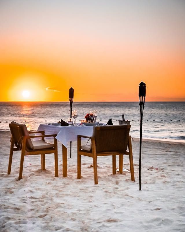 A romantic dinner table set up on a sandy beach with two chairs, torches, and a vibrant sunset over the ocean.
