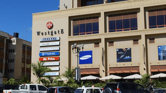 The large multistory shopping centre of Westgate Mall in Nairobi features various store signs on its exterior wall while several cars sit parked in the sunny lot out front.