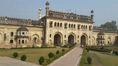 A wide-angle view of a majestic palace-like building with a grand arched entrance, multiple domes, and a lush green lawn in the foreground.