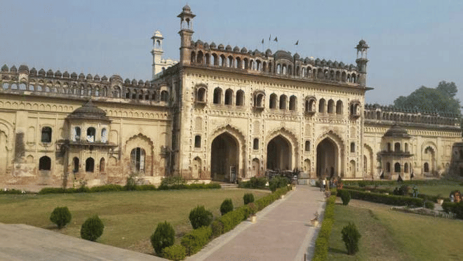 A wide-angle view of a majestic palace-like building with a grand arched entrance, multiple domes, and a lush green lawn in the foreground.