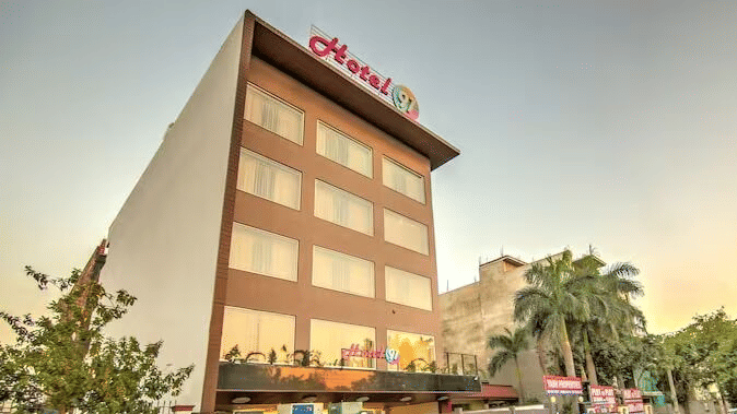 An exterior view of Hotel 91 HUDA Premier, Gurugram building with signage, entrance gate, parked car, and surrounding trees.