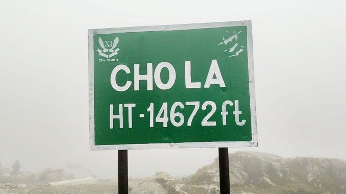 Green road sign at Cho La Pass in Sikkim, displaying altitude 14,672 ft, surrounded by misty high-altitude mountain landscape