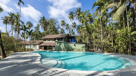 An outdoor swimming pool surrounded by a concrete deck, palm trees, and buildings at Oceanature Resort, Alibaug.