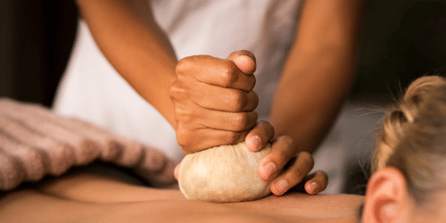 A pair of hands pressing a herbal pouch onto a person’s back lying on their stomach
