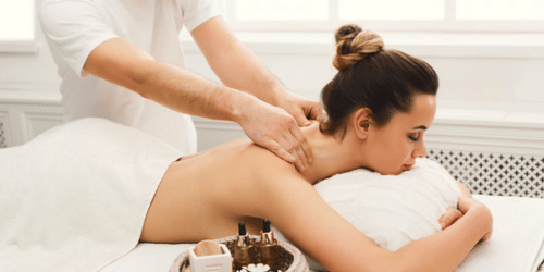 A woman lying on her stomach with her face resting on a pillow, receiving massage on her shoulders