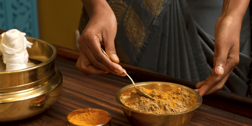A person holding a bowl of powdery substance in one hand and mixing it with the spoon on the other hand on a wooden surface next to a container with poultice at YO1 Longevity & Health Resorts