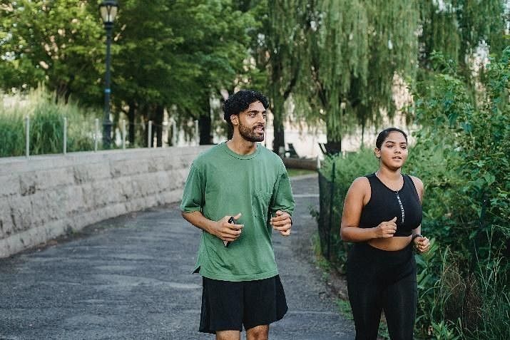 A man and woman jog together along a tree-lined path in a park, enjoying an active morning surrounded by lush greenery.