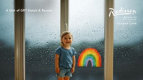 Creative artistic photography of a young child standing by rain-streaked windows holding a colorful rainbow decoration, capturing the joy and wonder of monsoon season during a family stay, promoting the Monsoon City Breaks package.