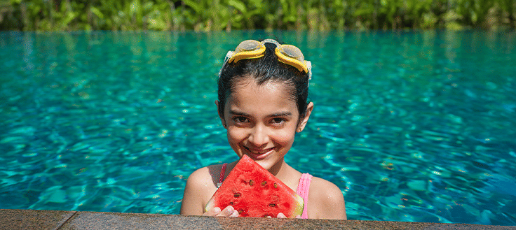 A young girl holding a slice of watermelon while standing in a swimming pool at Athiva Hotels & Resorts.