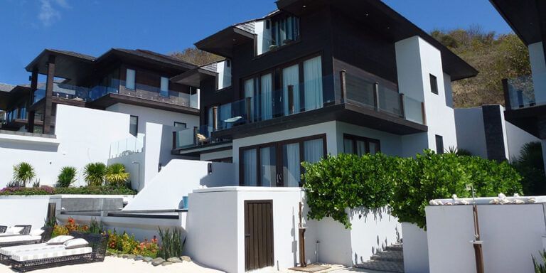 Modern beachfront villa with white walls and dark wood accents, featuring large glass windows, private balconies, and a landscaped outdoor area with sun loungers and greenery - Tamarind Hills Resort and Villas