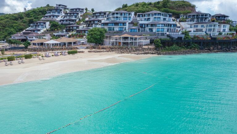 A picturesque beach scene features turquoise water, white sand, and a town built on a hillside overlooking the ocean.