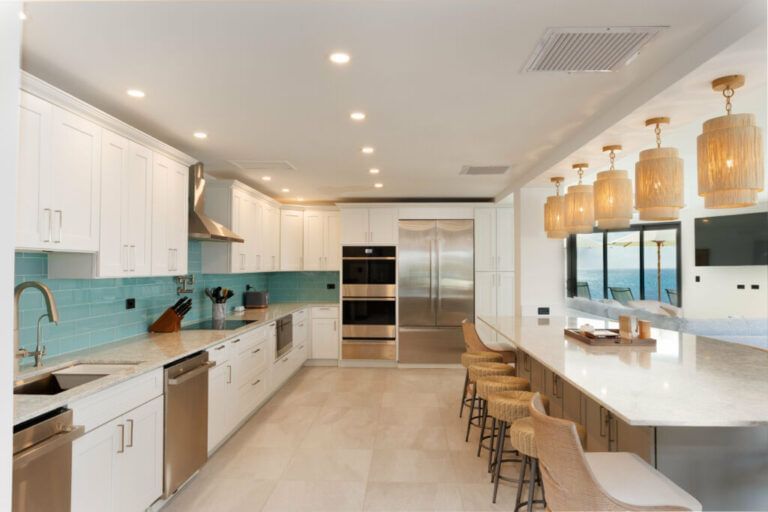 Spacious modern kitchen with white cabinetry, stainless steel appliances, turquoise backsplash, and a large island with wicker bar stools under pendant lights, overlooking an ocean view through large windows - Tamarind Hills Resort and Villas