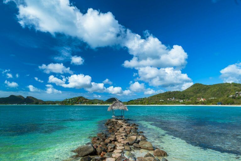 A scenic rocky path extends into crystal-clear turquoise waters, ending at a small thatched hut, with lush green hills and a bright blue sky with fluffy white clouds in the background.