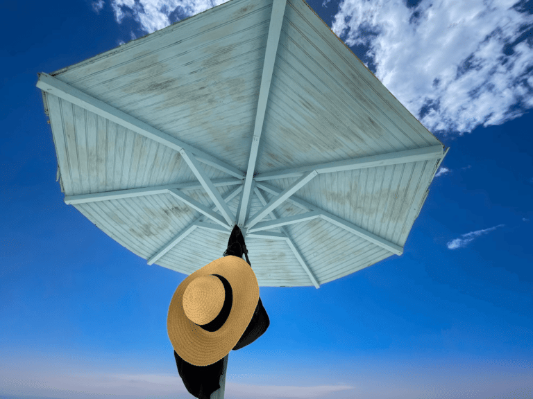 A wide-brimmed straw hat hangs from the center pole of a light blue wooden beach umbrella against a bright blue sky.