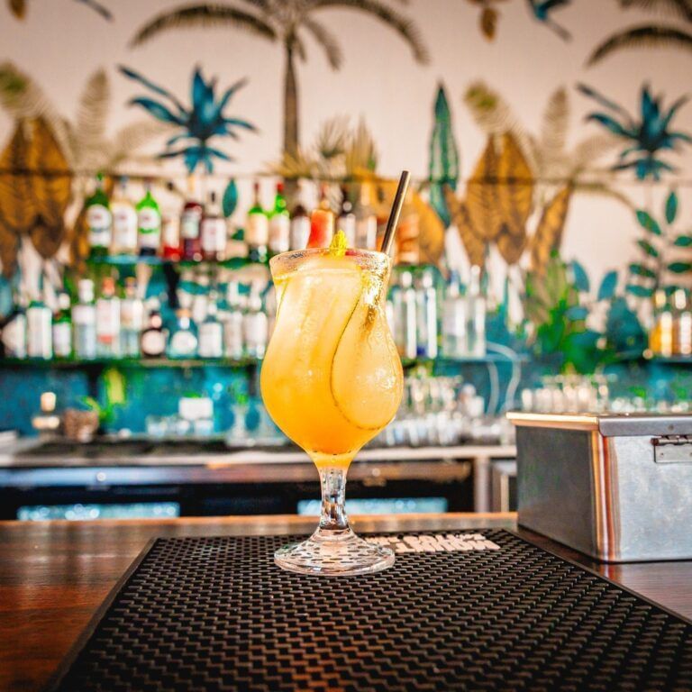 A colorful, garnished cocktail sits on a bar mat, with a vibrant mural of palm trees and a well-stocked bar in the background.