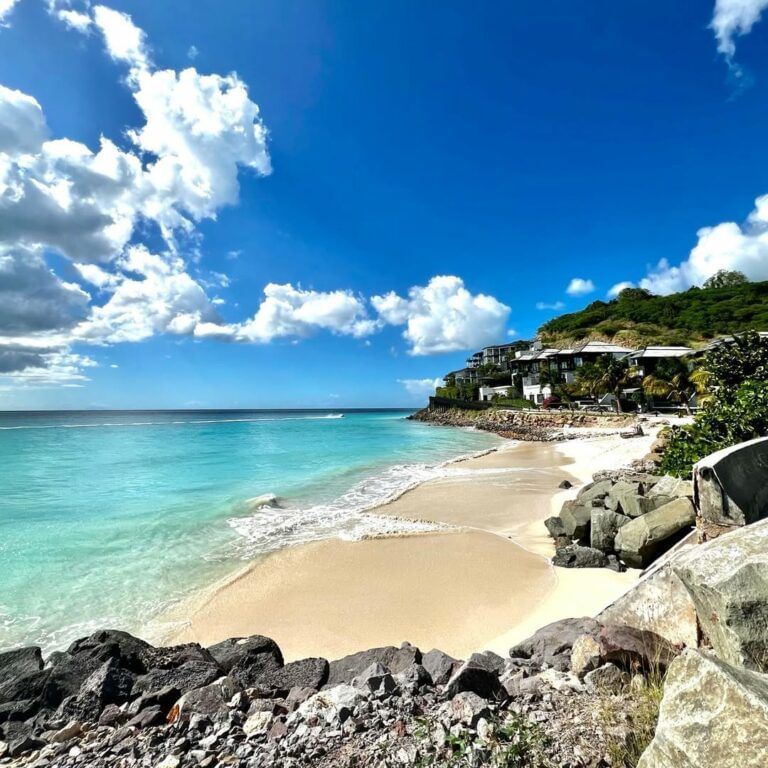 A picturesque bay with turquoise water, a sandy beach with large rocks, and resort buildings nestled on a green hillside.