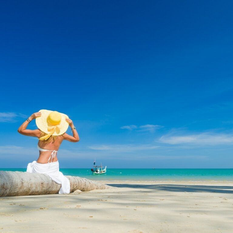 A woman in a white sarong and yellow hat sits on a fallen palm tree, gazing at a boat on the turquoise ocean.