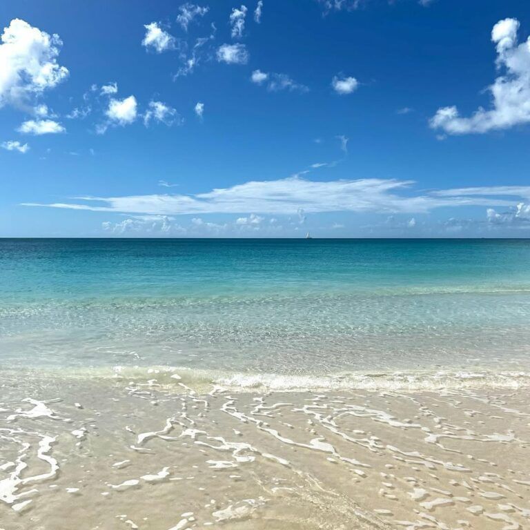 Clear, shallow turquoise ocean water gently laps onto a light sandy beach under a bright blue sky with white clouds.