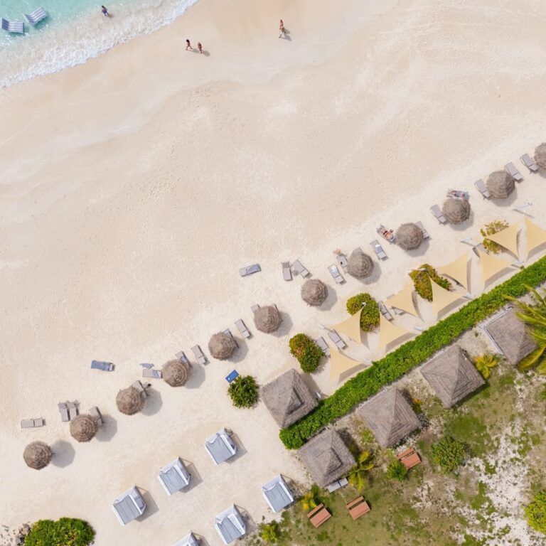 Aerial view of a tropical beach with soft white sand, rows of lounge chairs and thatched umbrellas, beachfront cabanas, and a few people walking along the shoreline by the turquoise water.