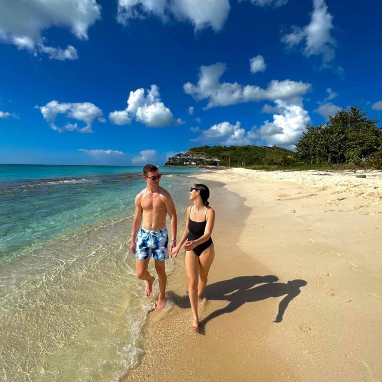 A couple walking along a sunny beach with clear blue water and sky.