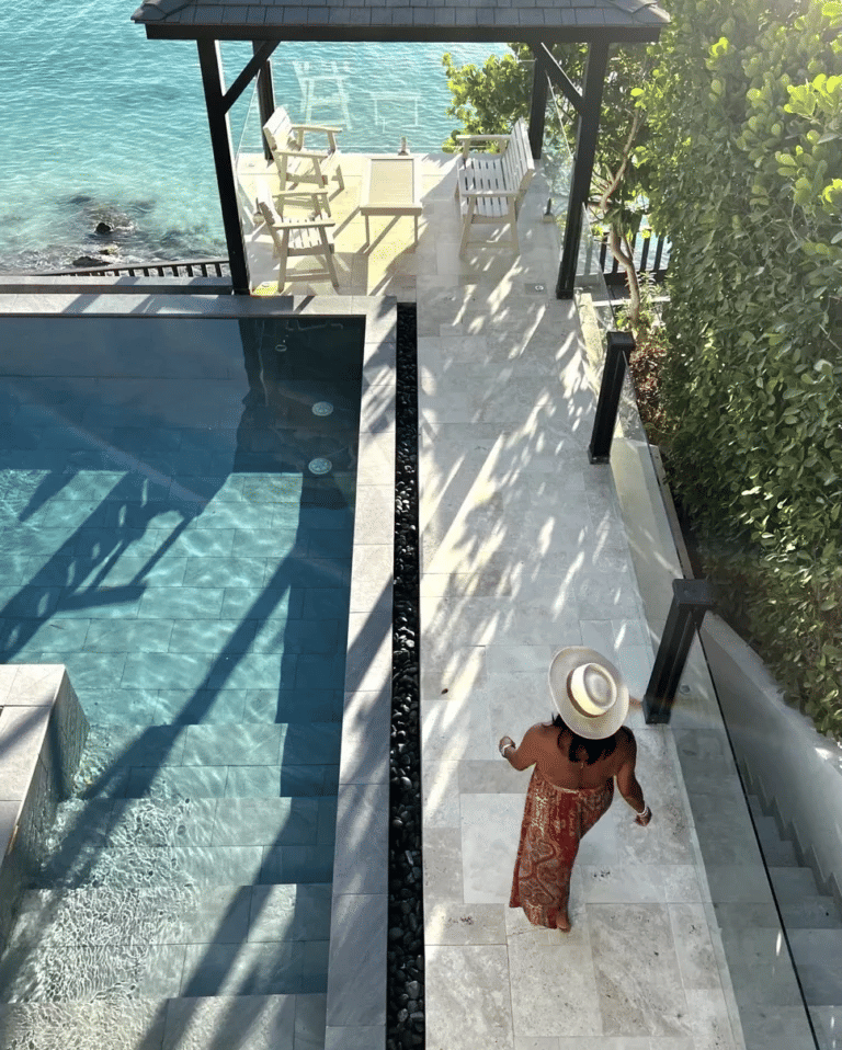 A person in a long skirt and hat walks down stone steps beside a bright pool, with the ocean and a shaded sitting area in view.