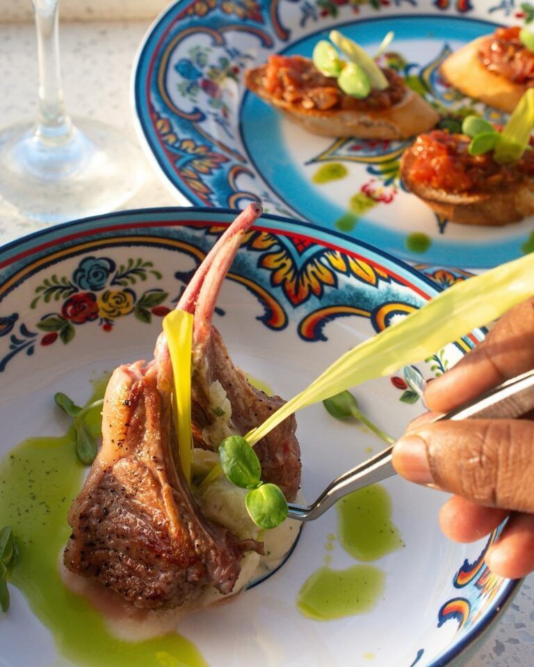 A chef garnishing a plate of lamb chops with microgreens and a vibrant green sauce on a decorative plate.