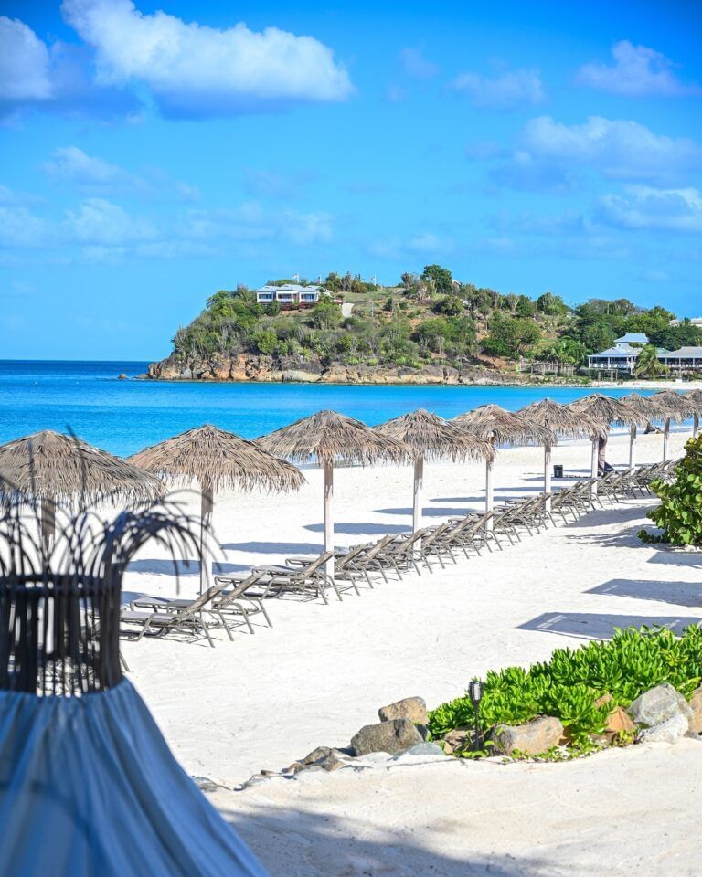 A long line of thatched umbrellas and lounge chairs stretch along a white sand beach with clear turquoise water and a green hillside.