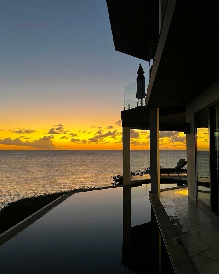 A luxury villa with an infinity pool at sunset, reflecting the vibrant orange and yellow hues of the sky over the calm ocean.
