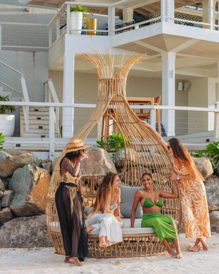 Four women socialize around a stylish, woven beach cabana on the sand, with a modern white building in the background.
