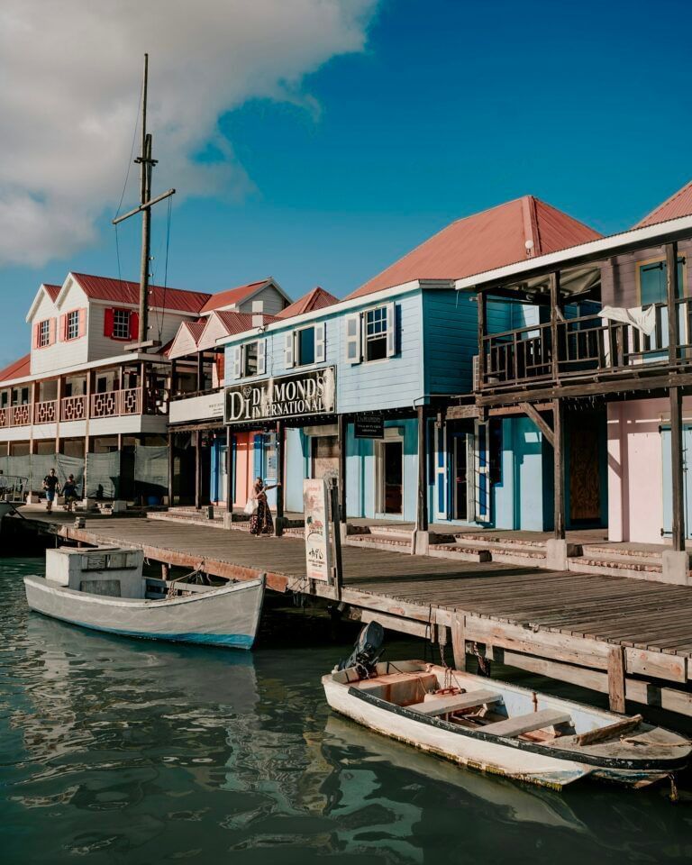 Colorful waterfront buildings with red roofs line a wooden boardwalk in a quaint Caribbean town, with small boats docked in the calm turquoise water and a few people strolling along the pier.