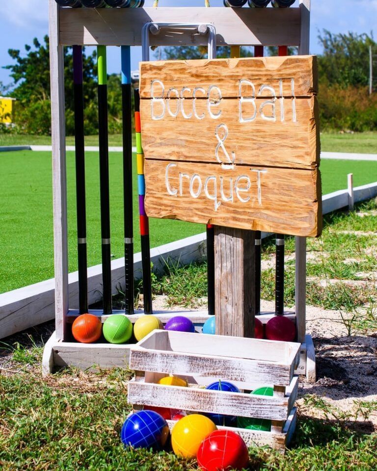 Outdoor bocce ball and croquet setup on a grassy area next to a green playing field, featuring colorful balls, mallets, and a rustic wooden sign labeled Bocce Ball & Croquet.