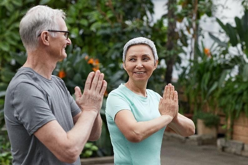 A smiling older couple practices yoga or meditation with hands pressed together, outdoors in a lush, green environment
