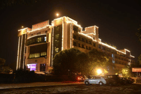 Night exterior of The Bristol Hotel, Gurgaon showing the multi-storey building outlined in string lights, the illuminated entrance, and cars parked in front.