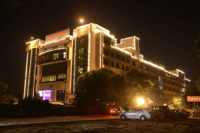 Night exterior of The Bristol Hotel, Gurgaon showing the multi-storey building outlined in string lights, the illuminated entrance, and cars parked in front.
