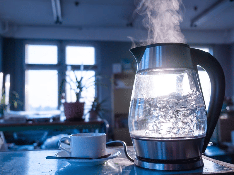 A steaming electric glass kettle boiling water beside a white cup and saucer on a table in a sunlit room, with blurred plants and shelves in the background.