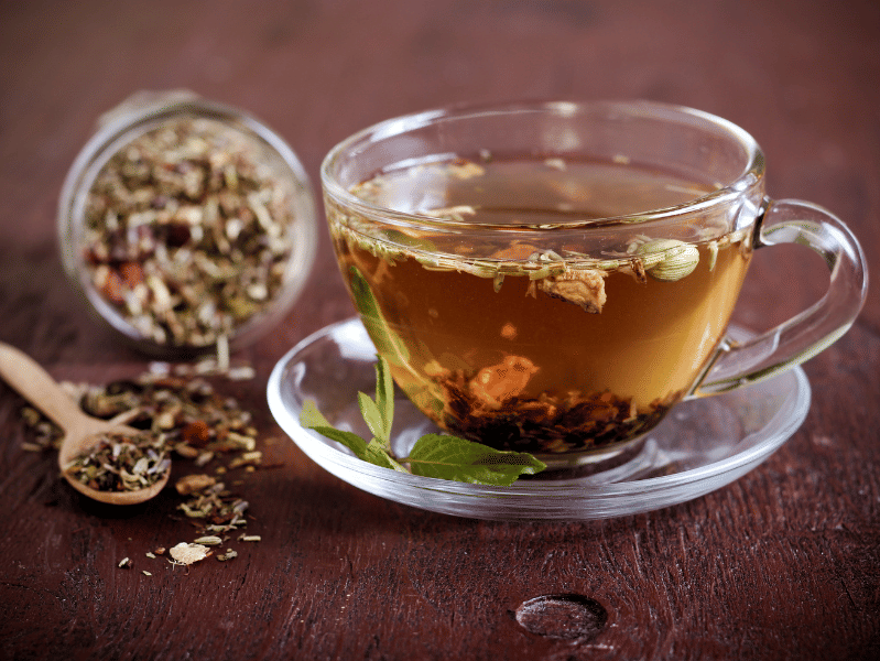 A glass cup filled with herbal tea, with loose-leaf tea floating inside and more loose-leaf tea spilled from a jar and wooden spoon on a wooden table.