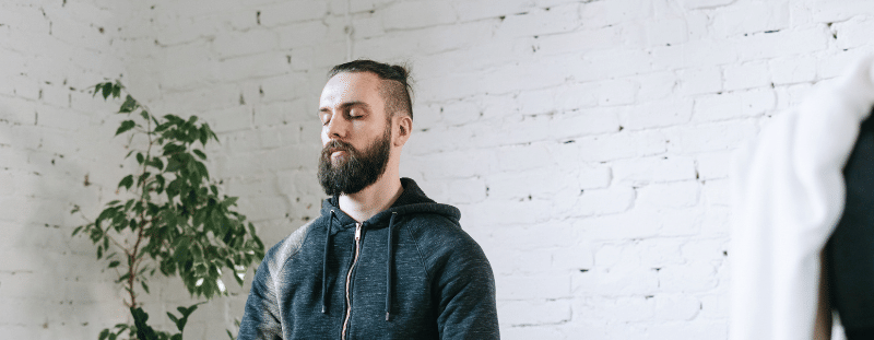 A person sitting cross-legged on a yoga mat with eyes closed, meditating in a calm indoor space with a plant and white brick wall in the background.