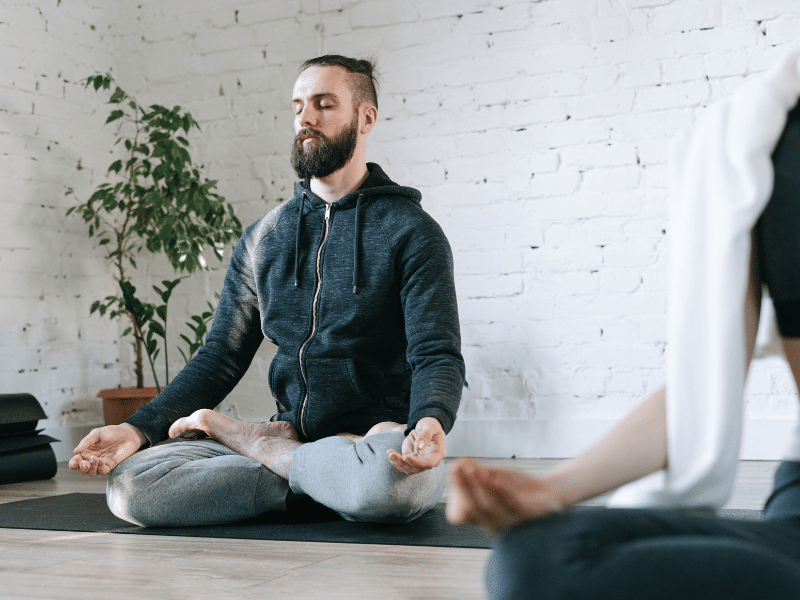 A person sitting cross-legged on a yoga mat with eyes closed, meditating in a calm indoor space with a plant and white brick wall in the background.