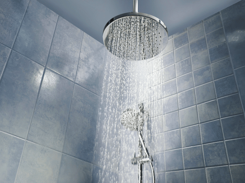 Water streaming from a modern round rainfall showerhead mounted on a tiled bathroom wall.