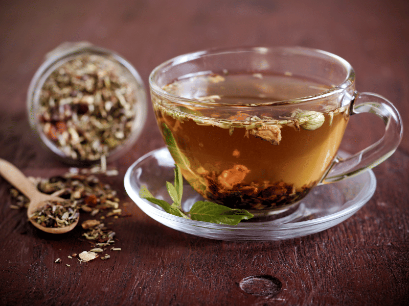 A glass cup filled with herbal tea, with loose-leaf tea floating inside and more loose-leaf tea spilled from a jar and wooden spoon on a wooden table.