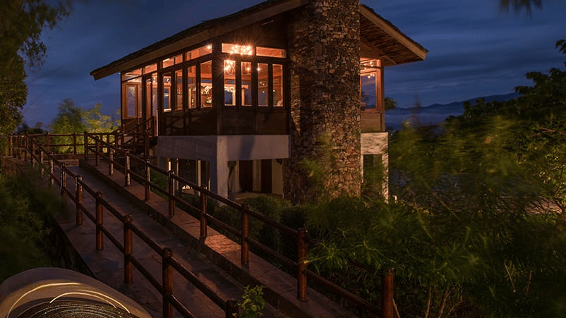 The exterior view of a raised stone-and-wood building with large windows, a walkway with railings, surrounding greenery, at The Serai Bandipur.