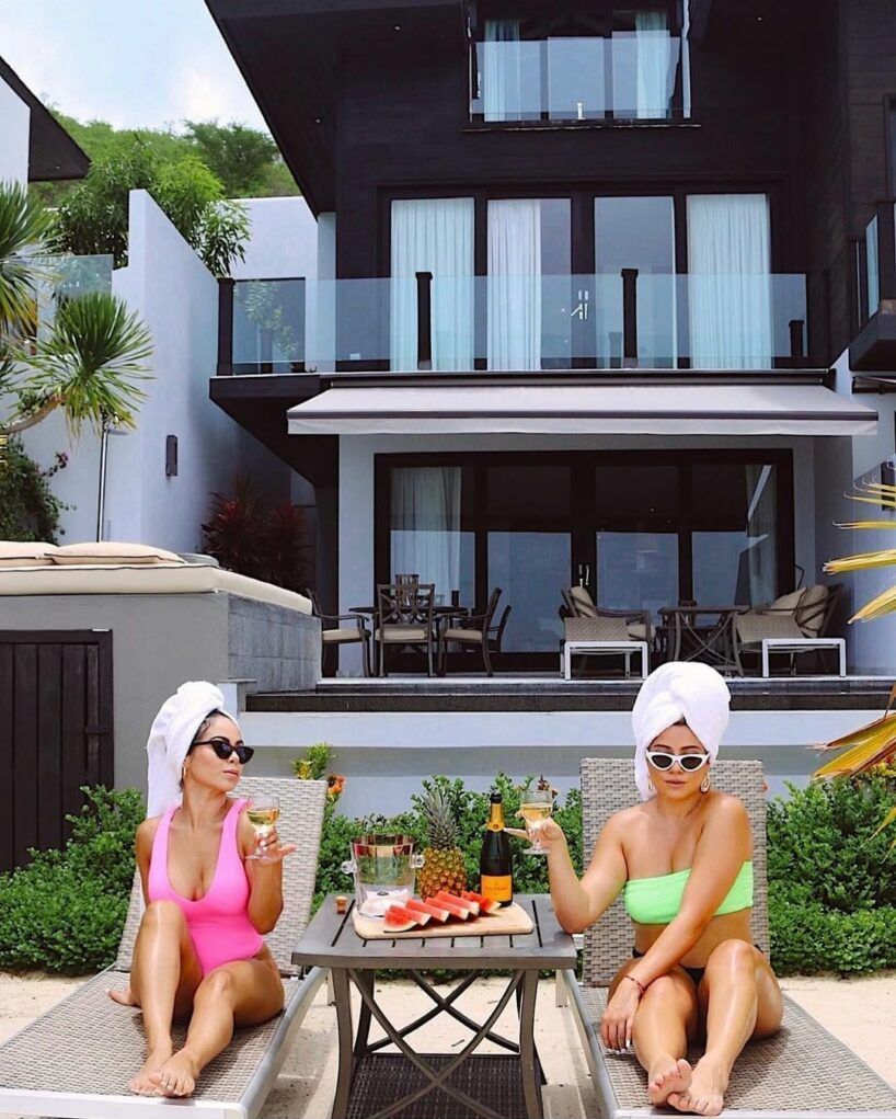 Two women in swimsuits and towels, with champagne and fruit, relaxing outside a modern villa.