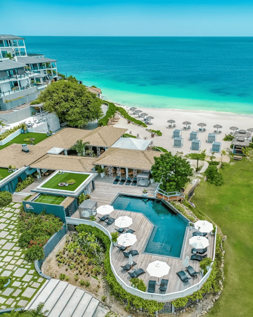 An aerial view of a luxurious resort complex with pools, villas, beach cabanas, and a white sand beach bordering the turquoise ocean.