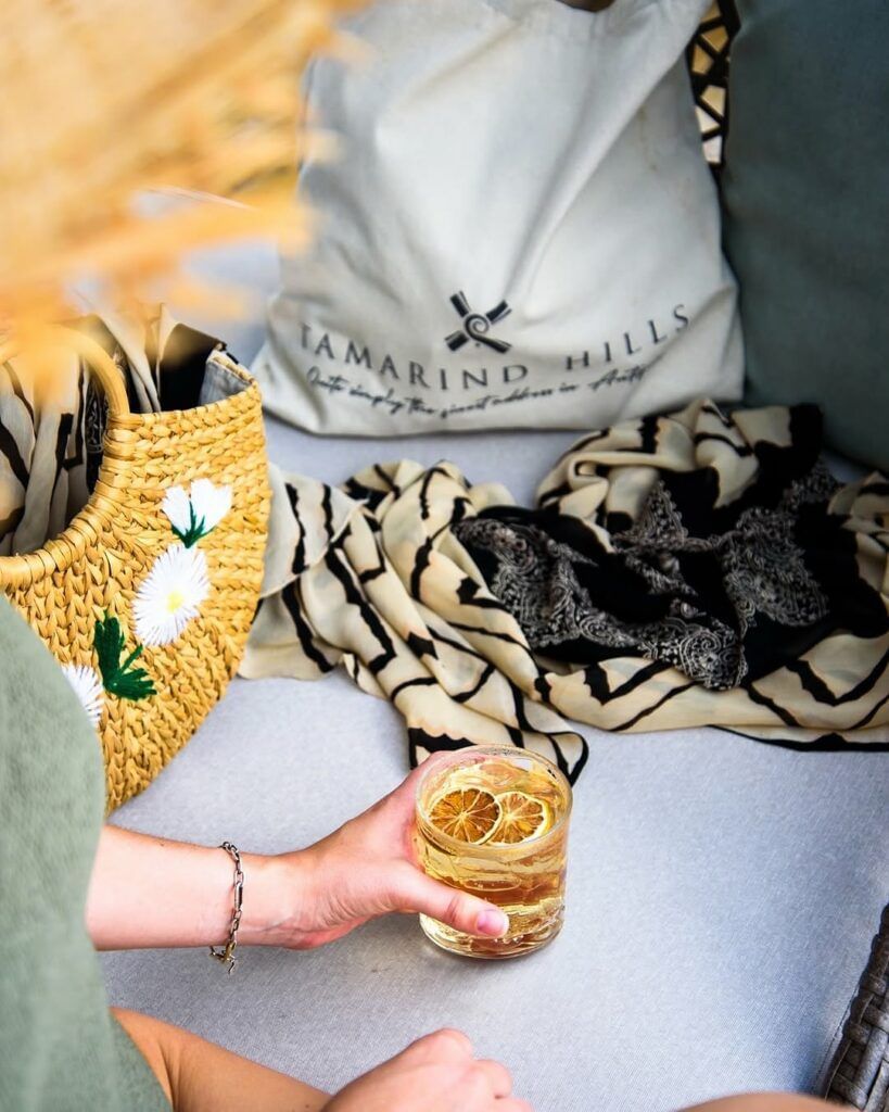 A person holding a drink with dried citrus while sitting on a couch next to a straw bag and a 'Tamarind Hills' tote.