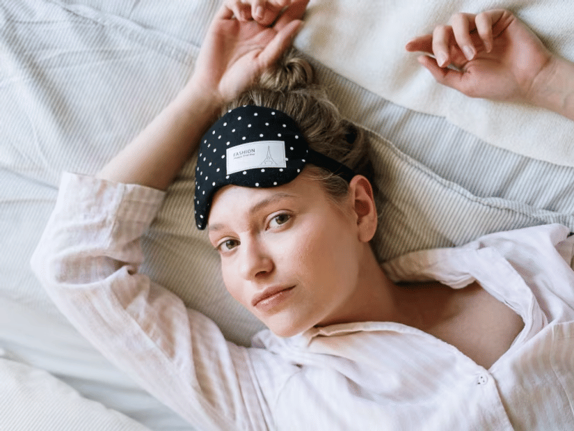 A woman lying in bed in pale pajamas with a polka-dot sleep mask pushed up on her forehead