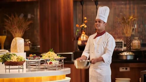 A chef in a white uniform and hat stands by a food counter, holding a bowl in a kitchen setting.