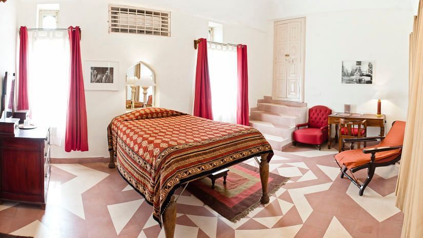 A room with a bed, chairs, a table, a dresser, patterned flooring, and a view into another room with stairs at Tijara Fort-Palace - 19th Century, Alwar.